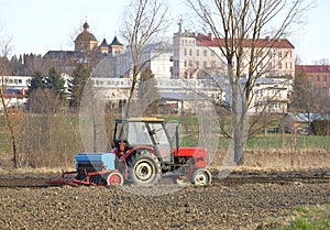 A farmer on a red tractor with a seeder sows grain in plowed land in a private field in the village area. Mechanization of spring