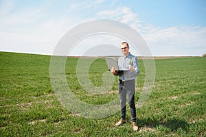 Farmer standing in young wheat field examining crop and looking at laptop