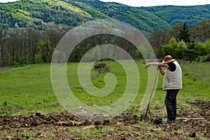 A farmer with a straw hat is taking a break after a hard work leaning on his shovel. He has just plowed the field on a spring day