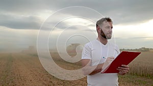 A farmer with a tablet for taking notes stands in a field next to combines working