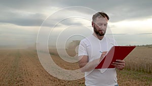 A farmer with a tablet for taking notes stands in a field next to combines working