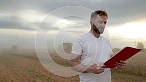 A farmer with a tablet for taking notes stands in a field next to combines working