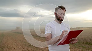 A farmer with a tablet for taking notes stands in a field next to combines working