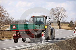 A farmer on a tractor pulls a trailer along an asphalt road passing among green fields. Rear view of multi-purpose agricultural
