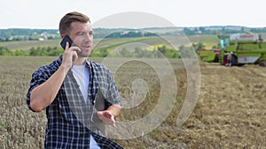 Farmer using a cell phone and holding notebook with a combine harvester in a wheat field on background