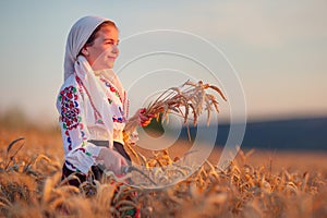 Farmer woman bulgarian little girl in ethnic folklore costume hold golden wheat straws and sickle in harvest field, harvesting and