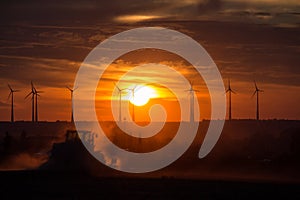 Farmers working with a tractor on the field at sunset with wind turbines in the background