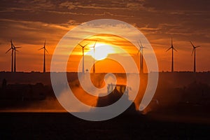 Farmers working with a tractor on the field at sunset with wind turbines in the background