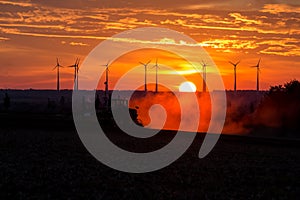 Farmers working with a tractor on the field at sunset with wind turbines in the background