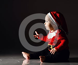 Fascinated baby boy toddler in red santa claus costume is sitting on ice looking carefully at copy space holding hand up