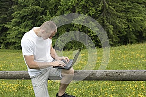 Fascinated man sitting on a wooden fence and works behind the laptop near the fields and pine forests