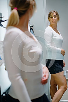 Fascinating lady stand in front of mirror in wardrobe, turning sideways checking new white longsleeve in reflection.