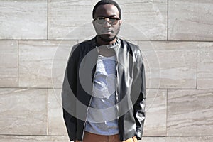 Fashion african man wearing a sunglasses and black rock leather jacket over textured gray background in city