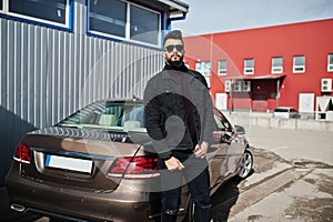 Fashion Arab man wear on black jeans jacket and sunglasses posed against business modern car. Stylish, succesful and fashionable