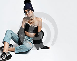 Fashion art photo of young grunge style girl in blue torn jeans, leather bralet and jacket, boots and winter hat sitting