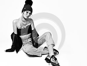 Fashion art photo of young grunge style woman in torn jeans, top, leather jacket, boots and winter hat sitting on floor