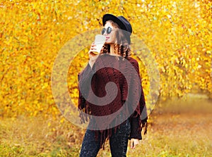 Fashion autumn style pretty young woman with coffee cup wearing a black hat sunglasses knitted poncho over yellow leaves