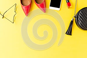 Fashion background. Red high heels, purse, phone, eyeglasses and nail polish on yellow table.