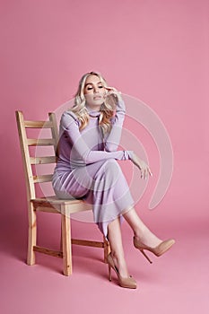 Fashion Beautiful blonde woman in knitted closed long dress sitting on a chair on a pink background. Perfect hair and makeup