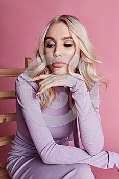 Fashion Beautiful blonde woman in knitted closed long dress sitting on a chair on a pink background. Perfect hair and makeup