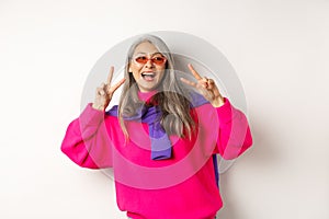 Fashion and beauty concept. Image of stylish asian senior woman in sunglasses smiling, showing peace signs and looking