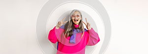 Fashion and beauty concept. Image of stylish asian senior woman in sunglasses smiling, showing peace signs and looking