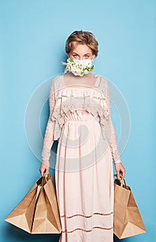 Fashion beauty cool young girl in medical mask designed with natural flowers standing with shopping bags in summer dress