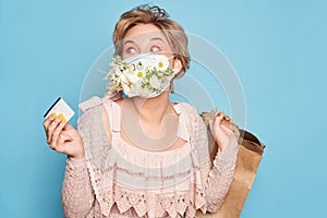 Fashion beauty cool young girl in medical mask designed with natural flowers standing with shopping bags in summer dress