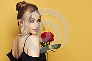Fashion beauty portrait of beautiful woman with blonde hairdo and makeup holds red rose flower on black background