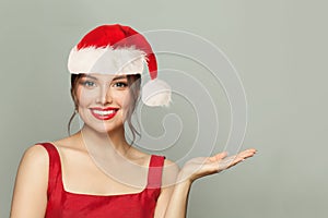 Fashion beauty portrait of Christmas woman. Beautiful female model in Santa hat posing with empty open hand on gray background