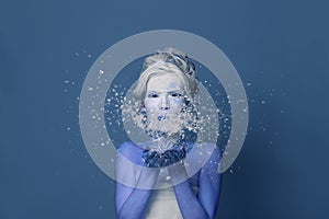 Fashion beauty portrait of winter Snow quuen woman with creative body art makeup posingblowing white snowflake on blue background