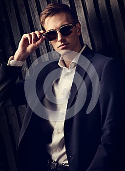 Fashion business man in suit posing in trendy eyeglasses on black studio wooden background. Closeup toned color elegant portrait