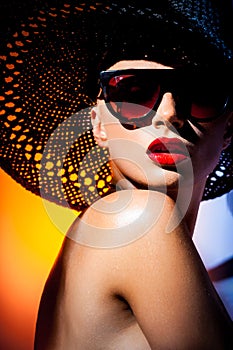 Fashion concept. Portrait of a beautiful woman in big hat and sunglasses in bright mixed contrast light. Female model