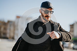 Stylish young man checking time on wristwatch while walking outdoors on sunny day in urban city area. photo