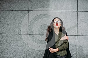 Fashion consept: beatiful young girl with long hair, glasses, red lips standing near modern wall wearing in green suit and grey je