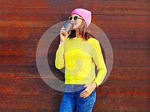 Fashion cool girl with coffee cup in colorful clothes over wooden background wearing pink hat yellow sweater