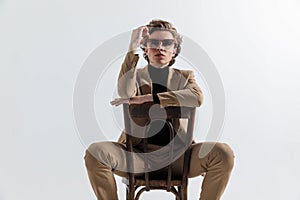 Fashion cool guy in smart casual outfit holding arms on chair and fixing glasses