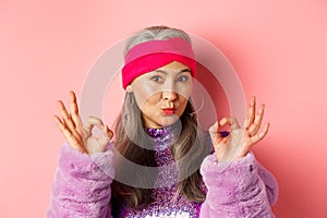 Fashion. Coquettish asian senior woman in trendy headband and faux fur coat, showing okay signs and pucker lips in kiss
