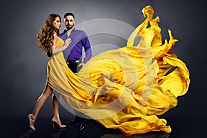 Fashion Couple Dancing. Woman in long Dress and Elegant Handsome Man. Lady in Yellow Silk Gown Flying on Wind over dark Gray