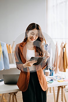 Fashion designer woman talking smart phone and using laptop with digital tablet computer in modern studio the clothes hanging on