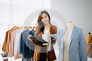 Fashion designer woman talking smart phone and using laptop with digital tablet computer in modern studio the clothes hanging on