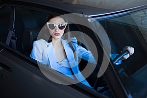 Fashion girl driving a car in a blue suit. Stylish woman sitting in the car and holding steering wheel