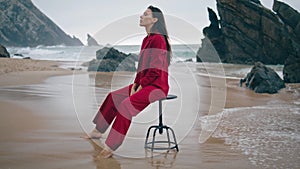 Fashion girl posing beach in red elegant suit overcast day. Woman sitting chair.