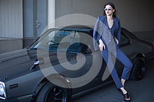 Fashion girl standing next to a retro sport car on the sun. Stylish woman in a gray suit waiting near classic car