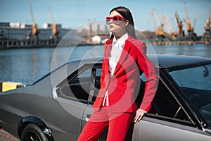 Fashion girl standing next to a retro sport car on the sun. Stylish woman in a red suit and sunglasses waiting near classic car