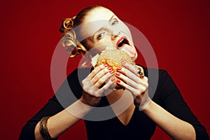 Fashion & Gluttony Concept. Happy red-haired model in black cocktail dress sticking out tongue & eating burger over red background
