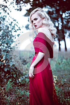 Fashion gorgeous young blonde woman in beautiful red dress in a fairy-tale forest. magic atmosphere. Retouched toning shot