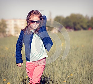 Fashion kid girl walking and posing in glasses and blue jeans jacket on spring sunny background