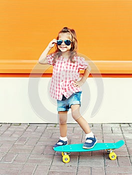 Fashion kid - smiling stylish little girl child with skateboard wearing sunglasses in city