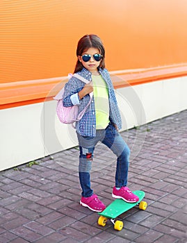 Fashion little girl child with skateboard wearing a sunglasses and checkered shirt and backpack over orange